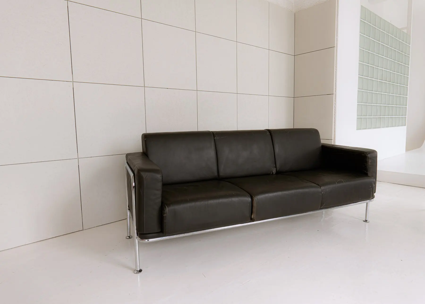 Melns ādas dīvāns hroma rāmī pie lielformāta flīžu sienas