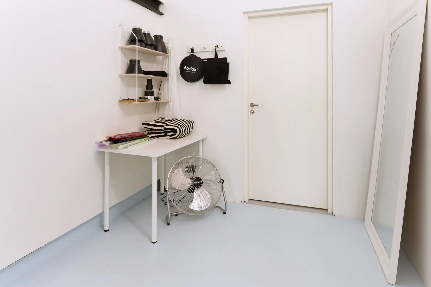 Plaukts ar reflektoriem un Godox softboksu, ventilators un liels stāvspogulis blakus zālēm