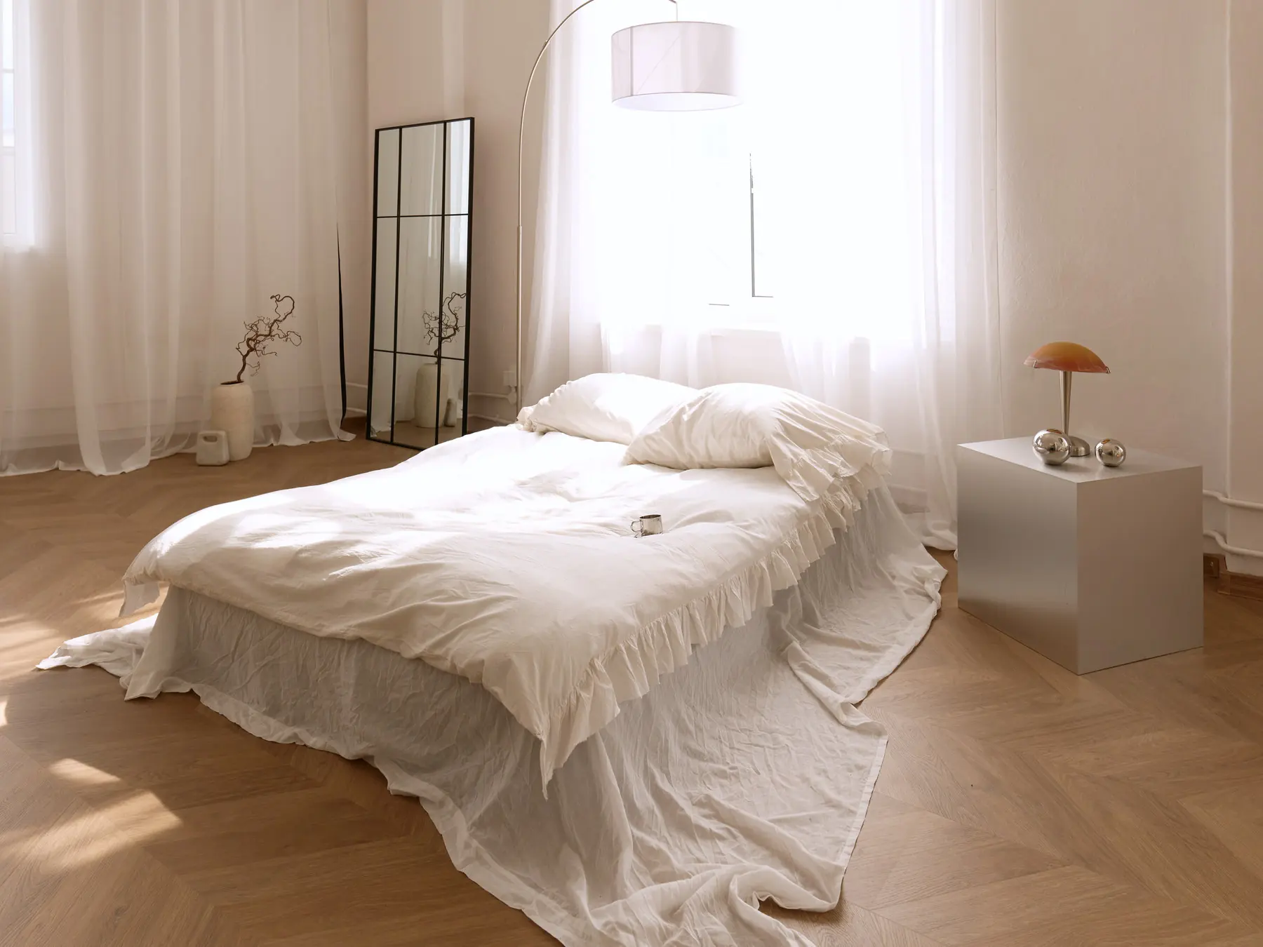 Home Chic zāle: gulta ar baltu gultasveļu, loka lampa un stāvspogulis pie gaišiem tilla aizkariem
